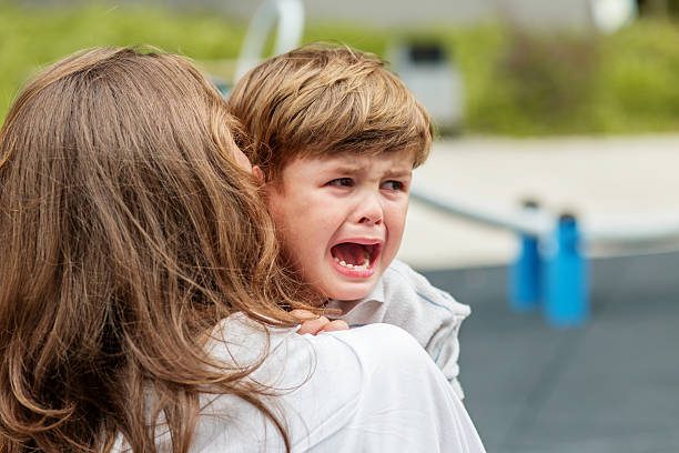 Upset little boy crying in his mother's arms at a playground, expressing distress and seeking comfort after emotional moment.